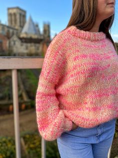 a woman wearing a pink and yellow sweater standing on a balcony with her hands in her pockets