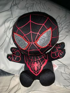 a spiderman stuffed animal sitting on top of a white bed sheet with black and red accents