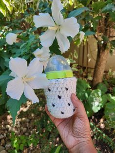 a hand holding a crocheted cup in front of white flowers and greenery