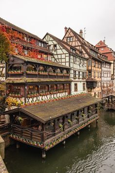 an old wooden building next to a river with lots of flowers growing on the roof