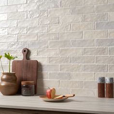 an apple on a plate next to two vases and a cutting board against a white brick wall