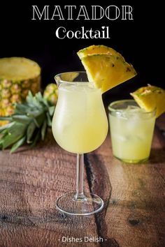 two glasses filled with drinks sitting on top of a wooden table next to pineapples