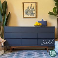 a blue dresser with lemons on top in front of a painting and potted plant