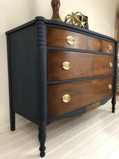 a black and brown chest of drawers with brass knobs on the top, against a white wall