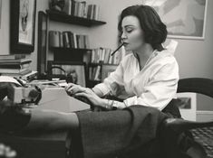 a black and white photo of a woman sitting at a desk with a pen in her mouth