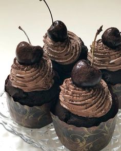 four cupcakes with chocolate frosting and cherries in a glass dish on a table