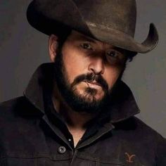 a man with a beard wearing a cowboy hat looking at the camera while standing in front of a gray background