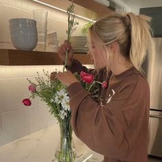 a woman arranging flowers in a vase on the kitchen counter top with shelves behind her