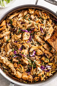 a skillet filled with chicken and vegetables on top of a white table next to a wooden spoon