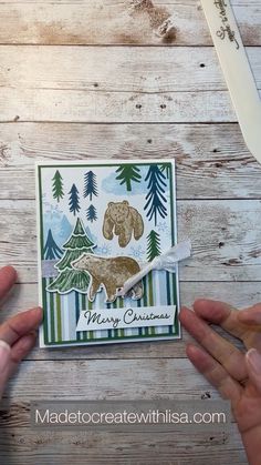 two hands holding up a christmas card with bear and pine trees in the background on a wooden table