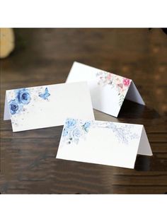 two cards with blue flowers on them sitting on a table next to a vase filled with flowers