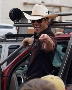 a man wearing a cowboy hat and sunglasses points at something in the distance while standing next to a car