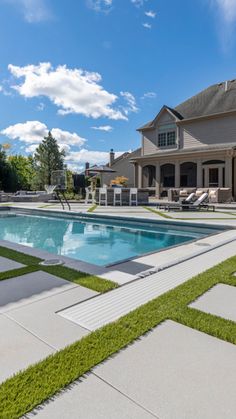 a pool surrounded by grass and lawn furniture in front of a house with a large swimming pool