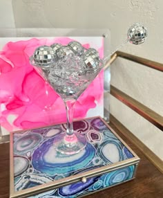 a martini glass sitting on top of a wooden table next to a mirror box and pink flower