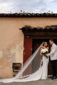 a bride and groom standing in front of a building with a veil blowing in the wind