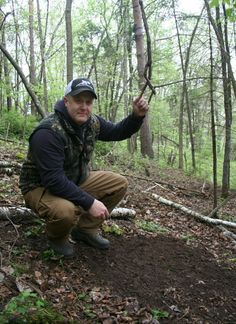 a man squatting down in the woods holding a branch with one hand and looking at the camera