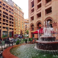 a fountain in the middle of a courtyard with people sitting at tables and chairs around it