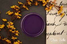 a purple paint can sitting on top of a table next to yellow flowers and an old letter