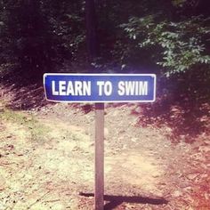 a blue sign that says learn to swim on the side of a dirt road in front of trees