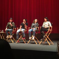 four people sitting on chairs in front of a red curtain, with one person speaking into a microphone