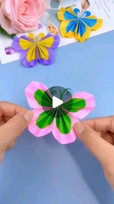 someone is holding a paper flower in front of some other flowers on a table with pink, yellow and blue colors