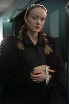 a woman in a black dress and fur coat with pearls on her head standing next to a wall