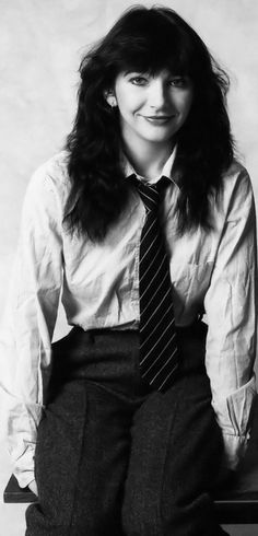 a woman sitting on top of a wooden table wearing a tie and dress pants with long hair