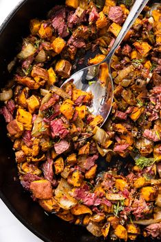 a skillet filled with meat and vegetables on top of a white countertop next to a silver spoon