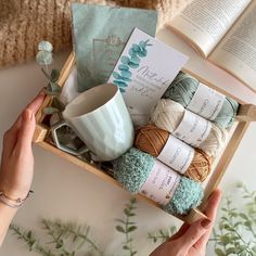 a person holding a box filled with yarn and crochet items next to an open book