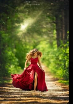 a woman in a long red dress walking down a dirt road with trees behind her