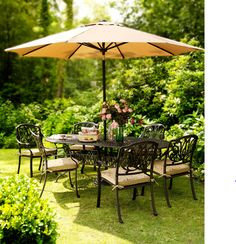 an outdoor dining table and chairs with umbrella in the middle of a lawn area surrounded by bushes