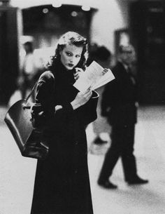 black and white photograph of a woman reading a newspaper while standing on the sidewalk in front of a train station