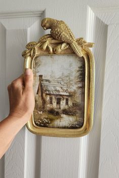 a person holding up a framed painting with a bird on it's head and a house in the background