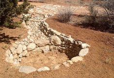 a stone wall in the middle of a dirt field with trees and bushes around it