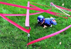 a small child in blue shirt and helmet crawling through pink tape on grass with red poles