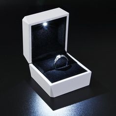 an open white box with a ring in it on a black surface and light shining through the lid