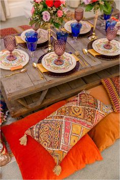 a wooden table topped with lots of colorful plates and place settings on top of pillows