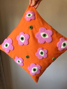 a hand holding an orange pillow with pink and green flowers on it, while someone is holding the pillow