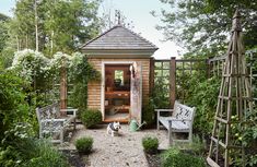 a small wooden shed in the middle of a garden with two chairs and a bench