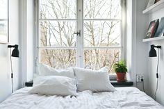 a bed with white sheets and pillows in front of a window that has a potted plant on it