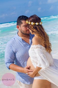 a pregnant couple kissing on the beach in front of the ocean with their belly wrapped around each other