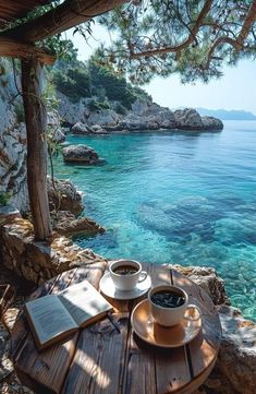 two cups of coffee sit on a wooden table overlooking the water's edge with an open book