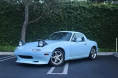 a light blue sports car parked in a parking lot next to a tall green hedge