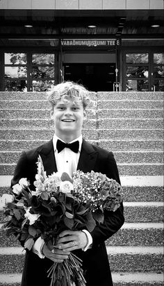 a boy in a tuxedo and bow tie holding flowers on the steps to a building