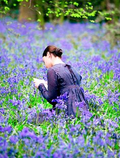 a woman sitting in the middle of a field with blue flowers on her knees, reading a book