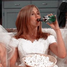 a woman sitting on a couch drinking from a bottle and eating popcorn in front of her