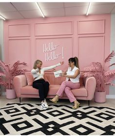 two women sitting on a pink couch in front of a wall with hello hello written on it