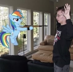 a young man standing in front of a window next to a rainbow colored pony on top of a couch