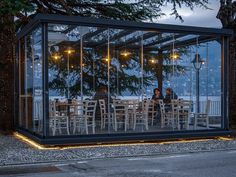 people are sitting at tables in a glass box on the side of the road with lights around them