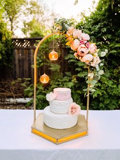 a three tiered wedding cake with pink flowers and greenery sits on a gold stand